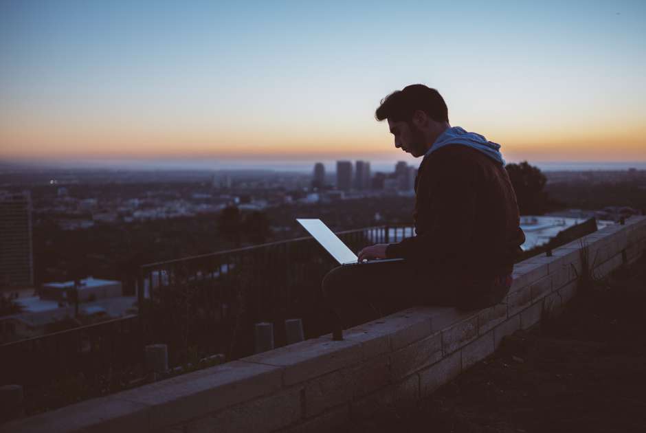 A man uses his computer while sitting on a building roof. (Avi Richards, Unsplash)